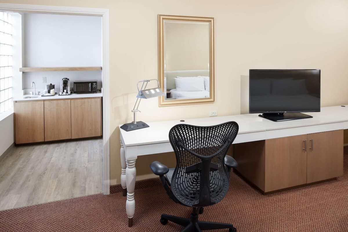 A hotel room with a white desk, a black office chair, a television, a mirror, and a kitchenette with a microwave and toaster.