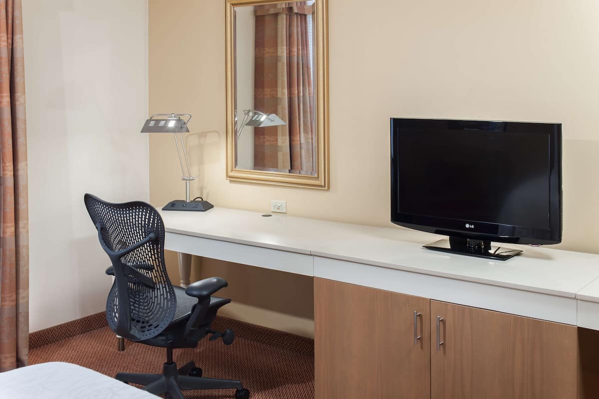A hotel room with a desk, a chair, a television, a lamp, and a mirror.