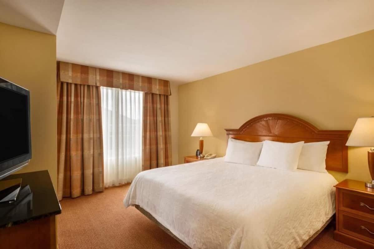 A hotel room with a bed, two bedside tables with lamps, a TV, and a window with curtains.
