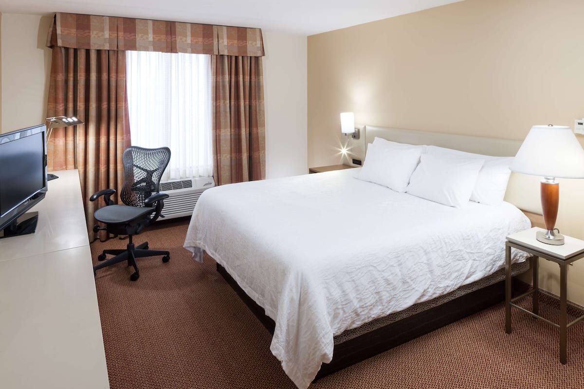 A hotel room with a large bed, a desk with a computer, a chair, a lamp, and a window with curtains.