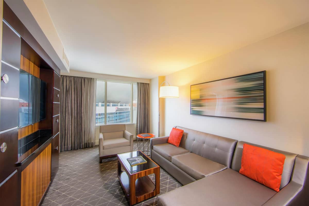 A hotel room with a sofa, armchair, and a small table. There is a TV on the left side and a window with curtains.