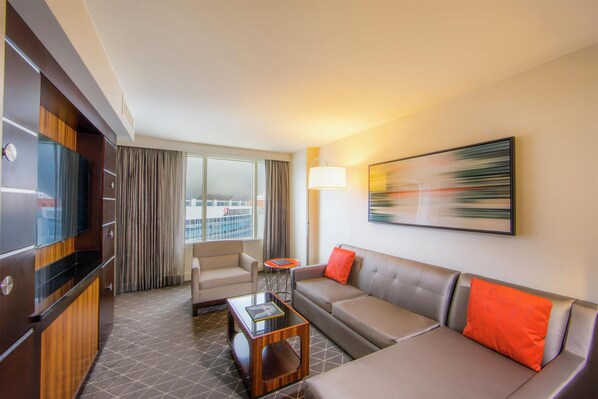 Habitación de hotel con sofá, sillón y una mesita. Hay un televisor a la izquierda y una ventana con cortinas.