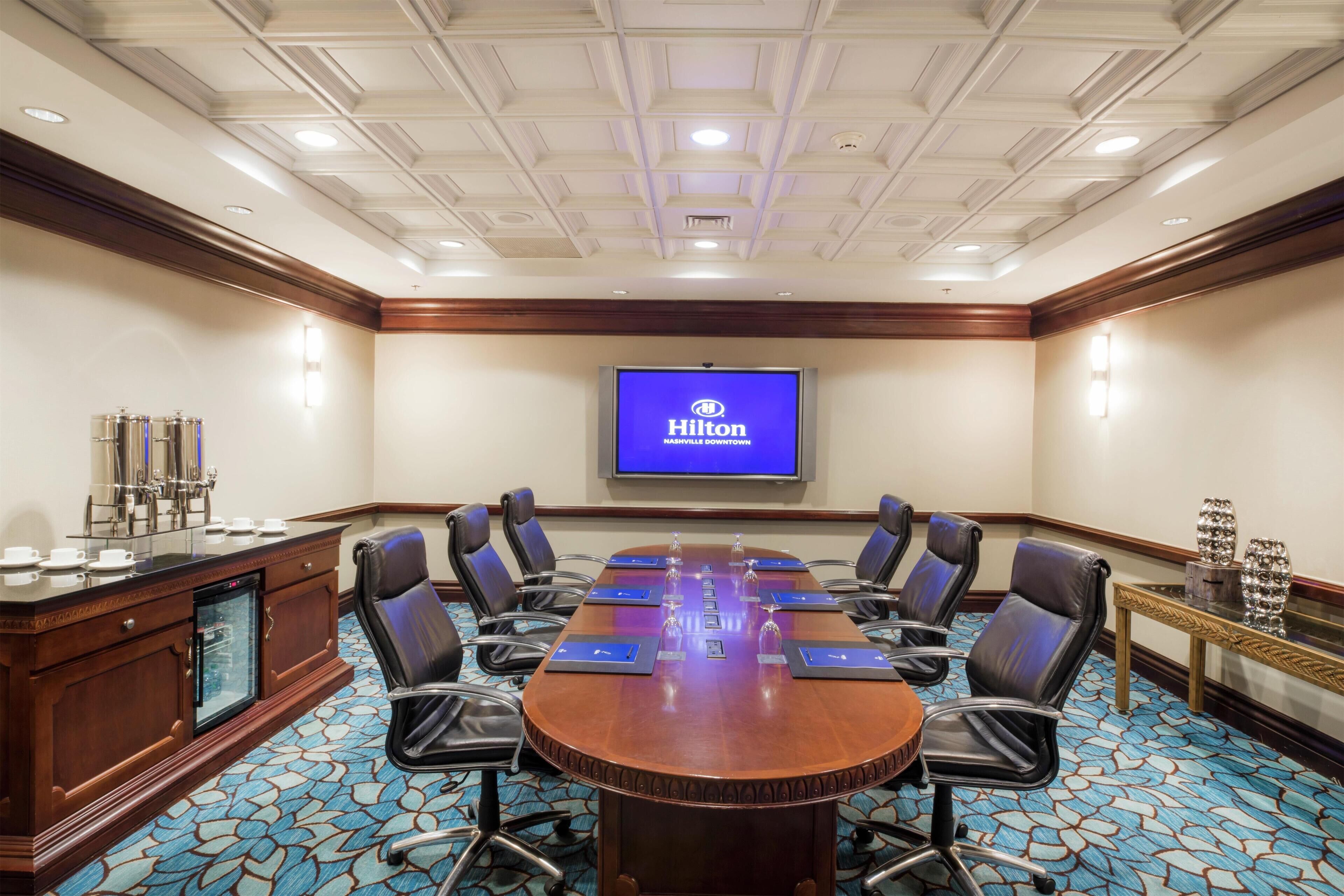A conference room with a long table, black chairs, a minibar, and a TV displaying the Hilton logo.