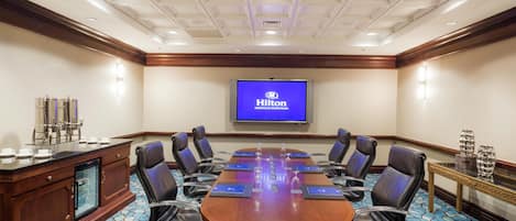 A conference room with a long table, black chairs, a minibar, and a TV displaying the Hilton logo.