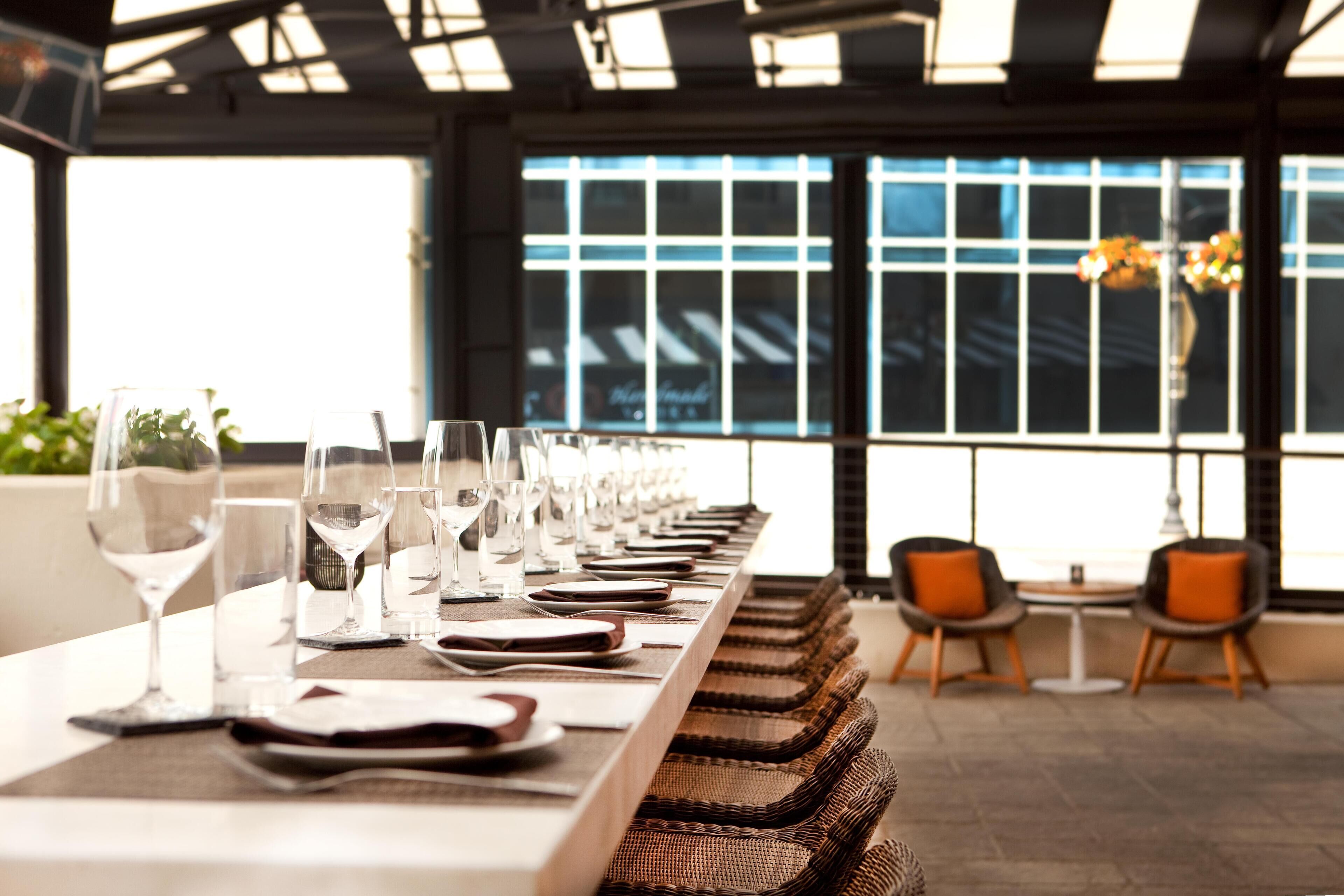 A long dining table with multiple wine glasses and plates, wicker chairs, and a view of the city through large windows.