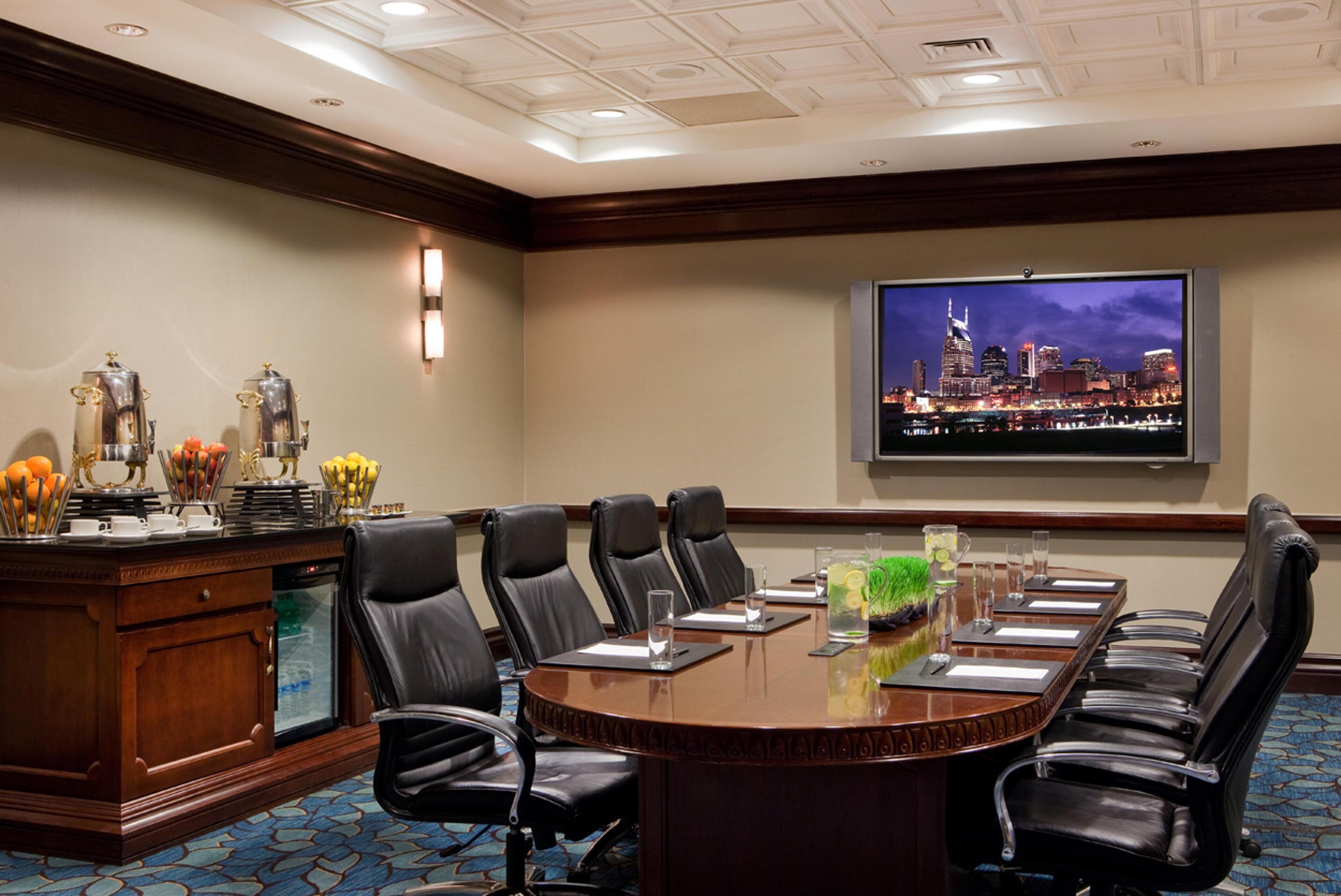 Un salón de conferencias con una gran mesa, varias sillas, un atril y una pantalla de televisión que muestra un paisaje urbano.