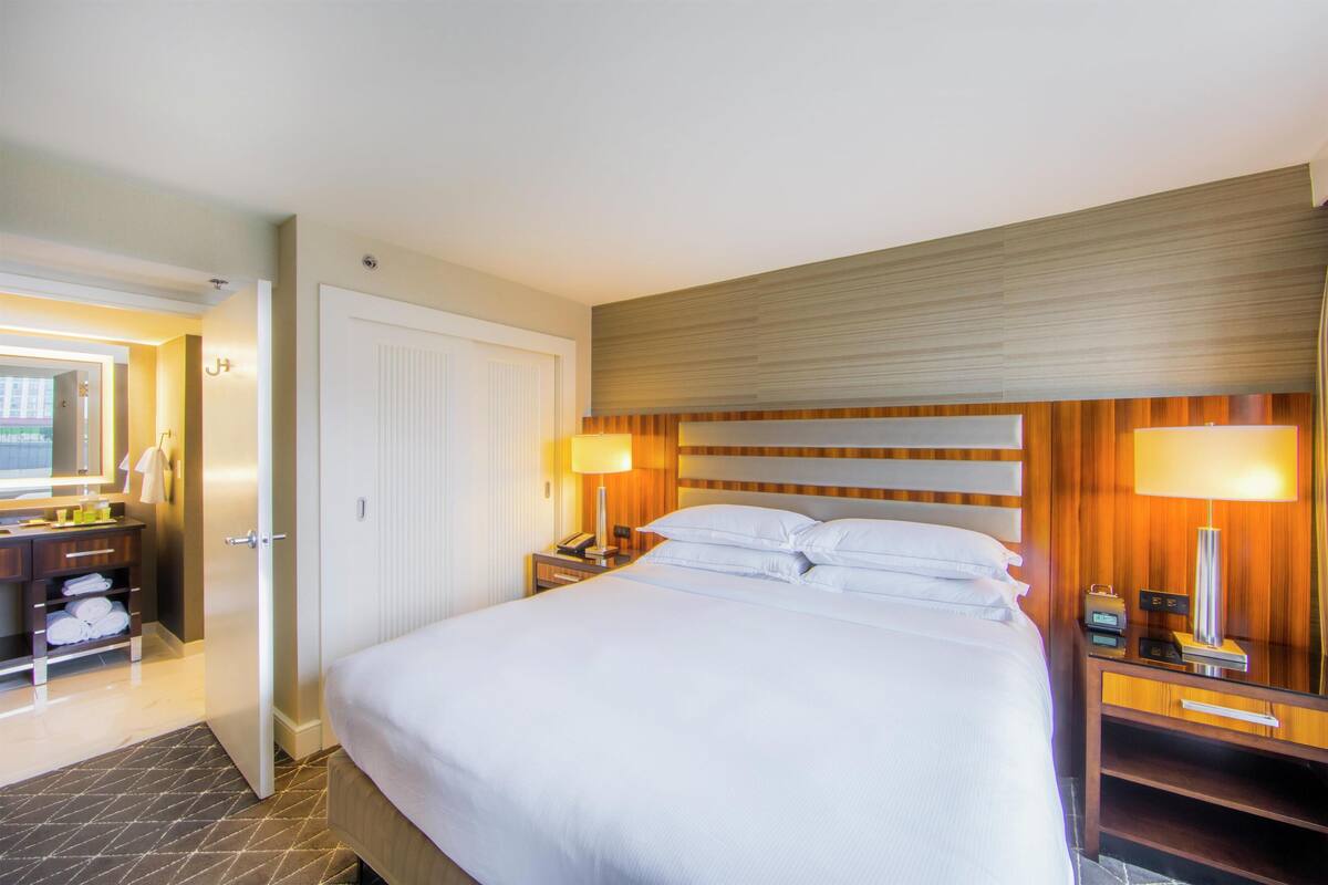 A hotel room with a large bed, bedside lamps, a wooden headboard, a nightstand, and a bathroom with a mirror and towel rack.