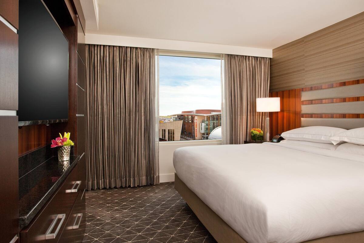 A hotel room with a large bed, a TV mounted on the wall, a window with curtains, and a view of buildings outside.