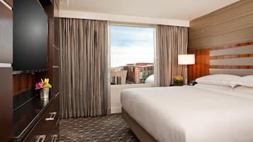Habitación de hotel con una cama grande, televisor montado en la pared, ventana con cortinas y vistas a edificios.