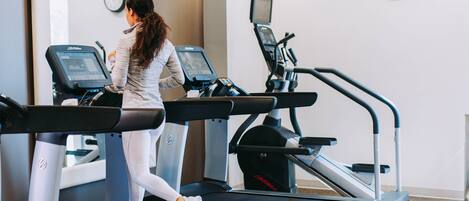 A woman running on a treadmill in a gym with multiple treadmills and a clock on the wall.