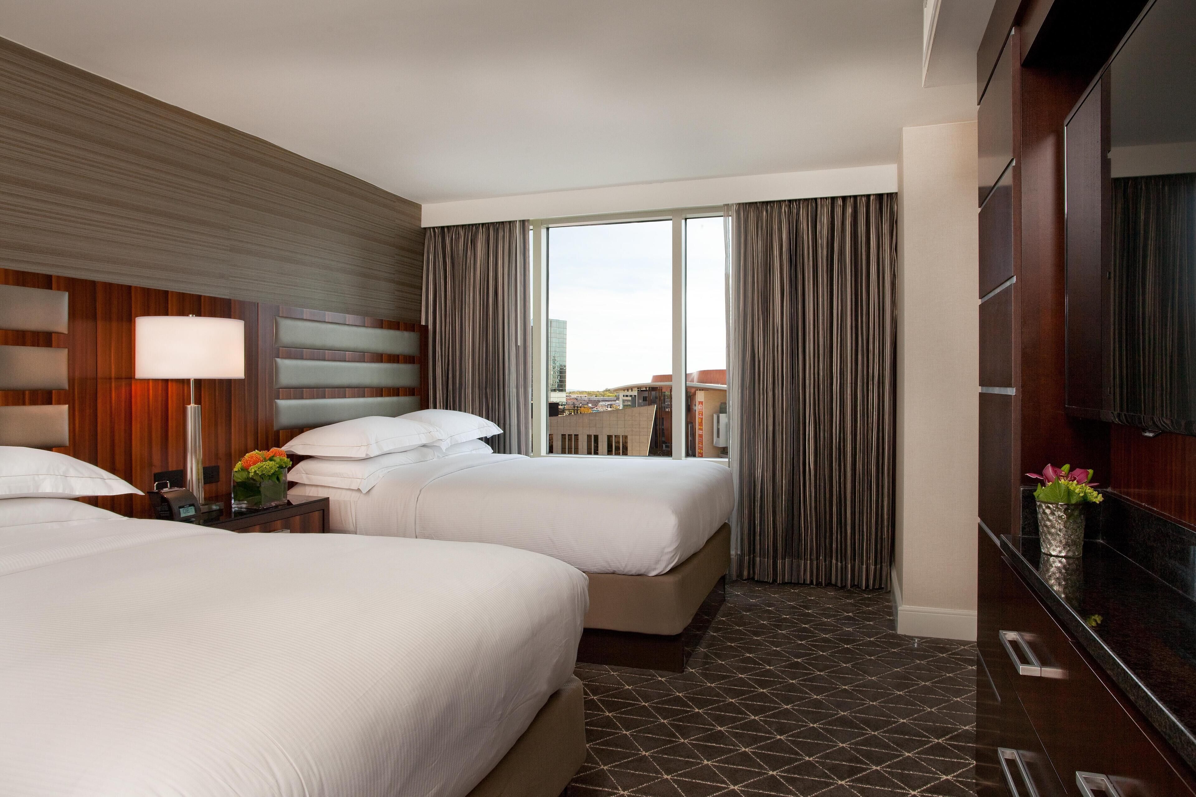 A hotel room with two beds, a large window with curtains, a TV, and a wooden wall panel.
