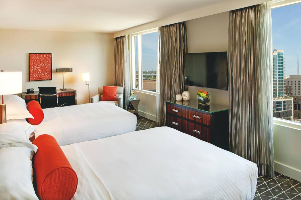 A hotel room with two beds, a TV, a desk, and a view of the city.