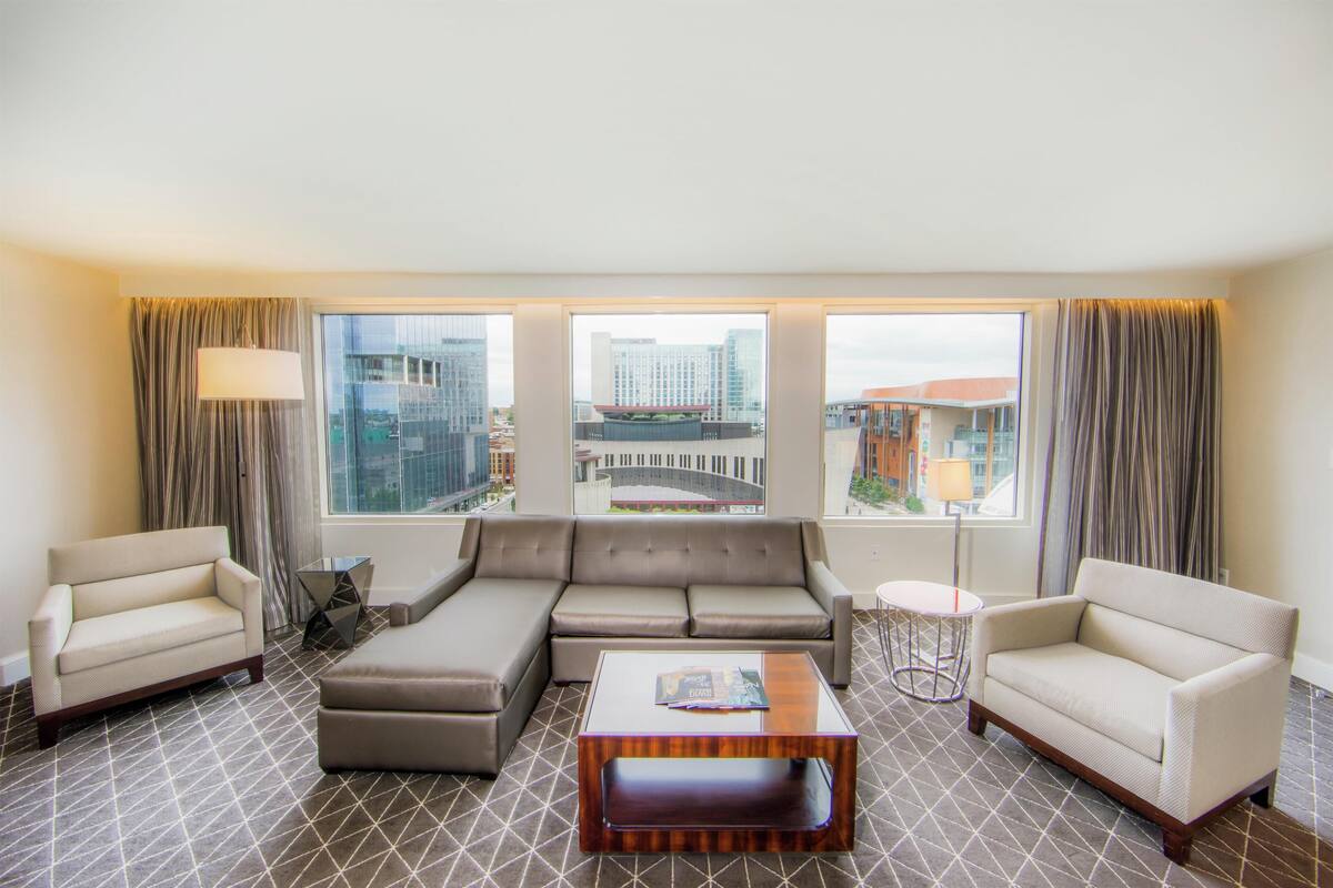 A modern hotel room with a large sectional sofa, two armchairs, a small round table, and a coffee table. The room features large windows with city views.