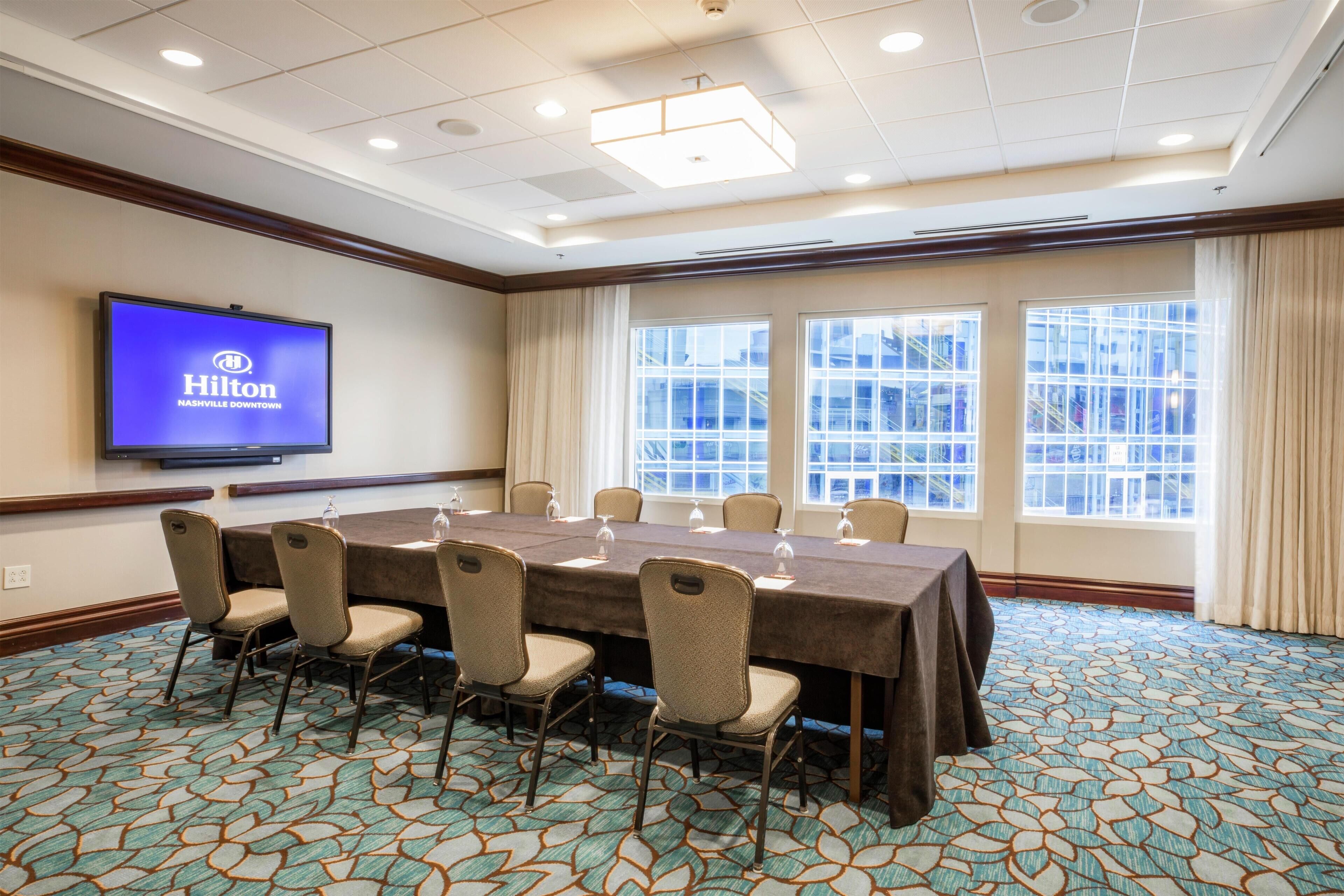 Un salón de conferencias con una mesa larga, sillas, una pantalla de televisión mostrando 'Hilton Nashville Downtown', ventanas amplias y alfombra estampada.