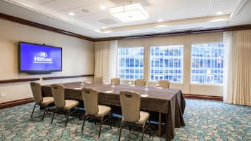 Un salón de conferencias con una mesa larga, sillas, una pantalla de televisión mostrando 'Hilton Nashville Downtown', ventanas amplias y alfombra estampada.