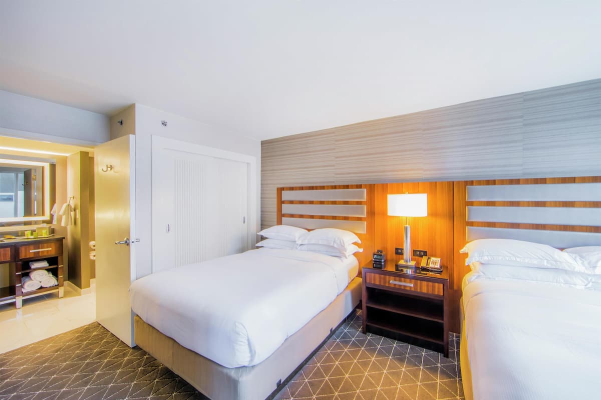 A hotel room with two beds, a bedside table with a lamp, a wardrobe, and a bathroom with a mirror and towel rack.