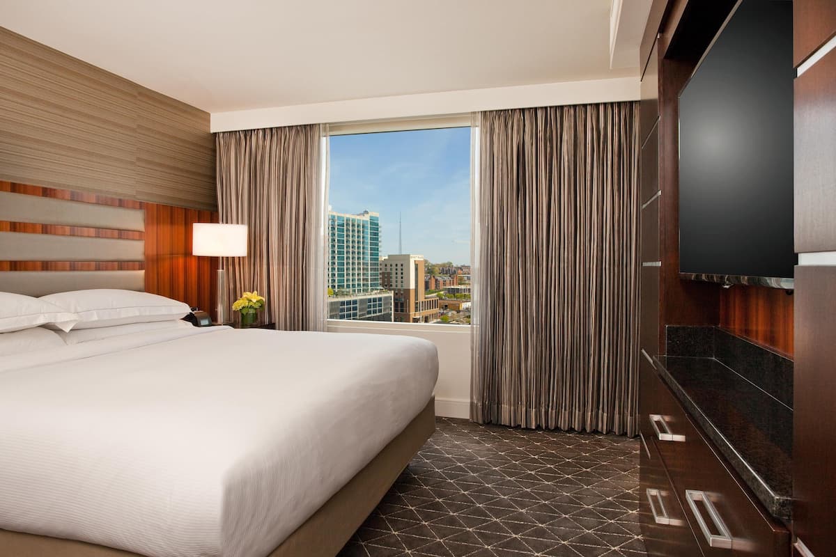 A hotel room with a large bed, a flat-screen TV, a window with curtains, and a city view.