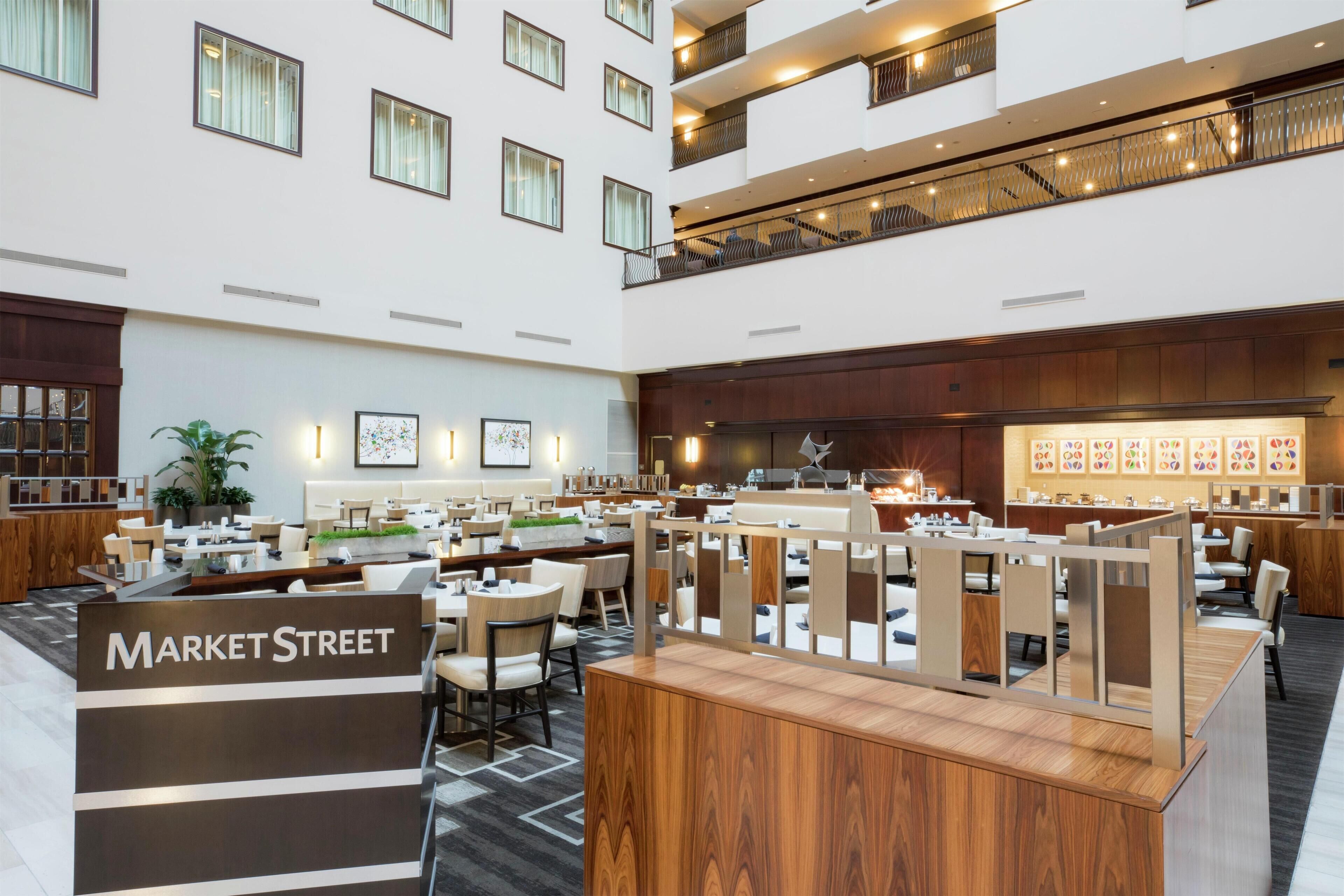 A spacious hotel lobby with multiple dining areas, a reception desk labeled 'Market Street', and a second-floor balcony with seating.