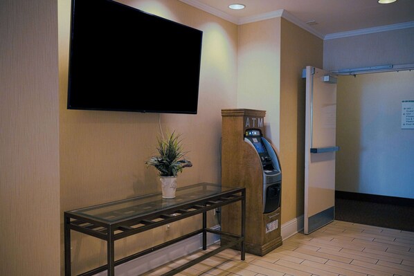 A reception area with a flat-screen TV mounted on the wall, a glass table with a potted plant, an ATM machine, and a door with a sign indicating guest services.