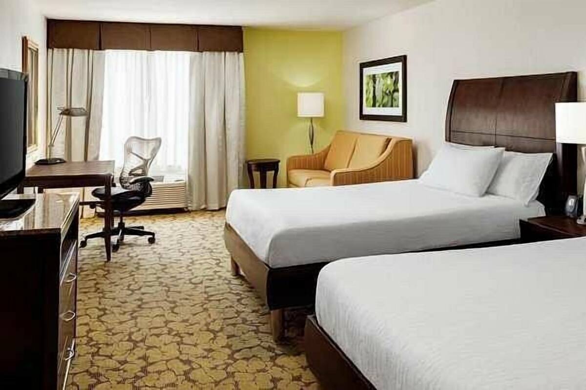 A hotel room with two beds, a desk, a chair, and a lamp.