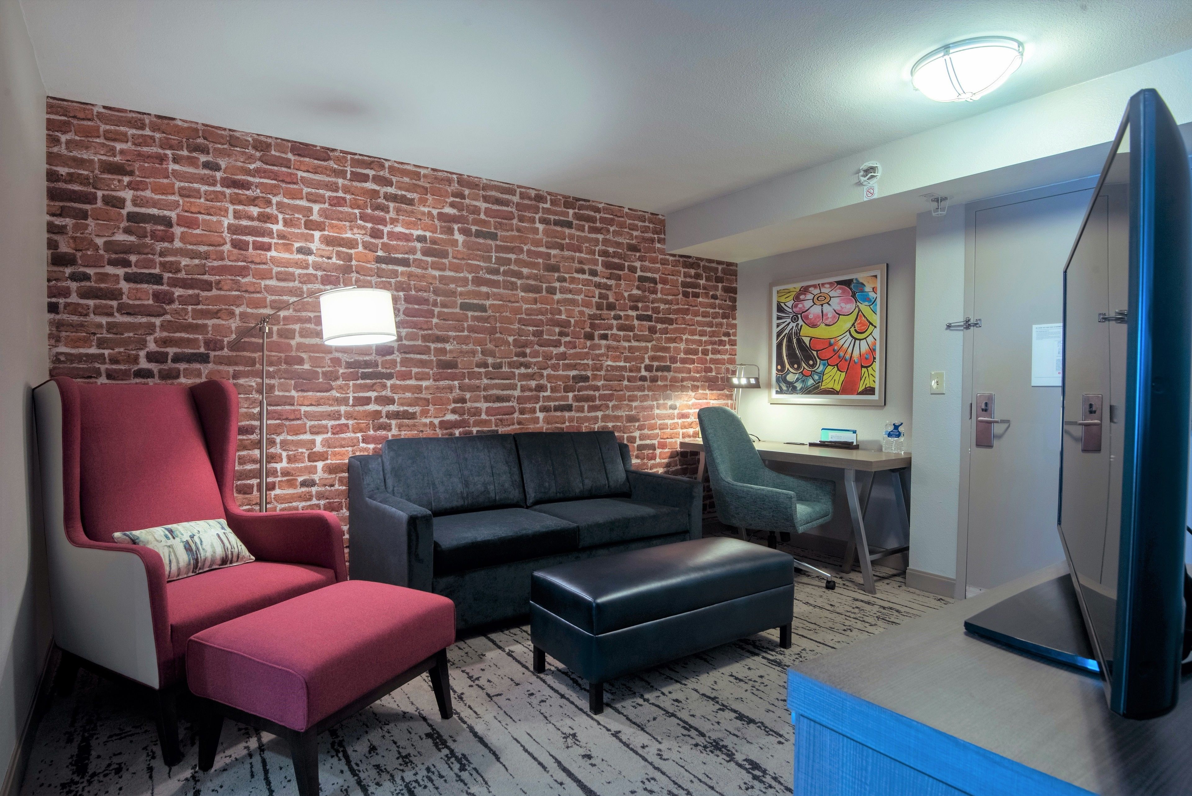 A room with a brick wall, a red armchair, a grey sofa, a black ottoman, a desk with a chair, and a TV.