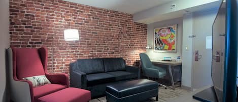 A room with a brick wall, a red armchair, a grey sofa, a black ottoman, a desk with a chair, and a TV.