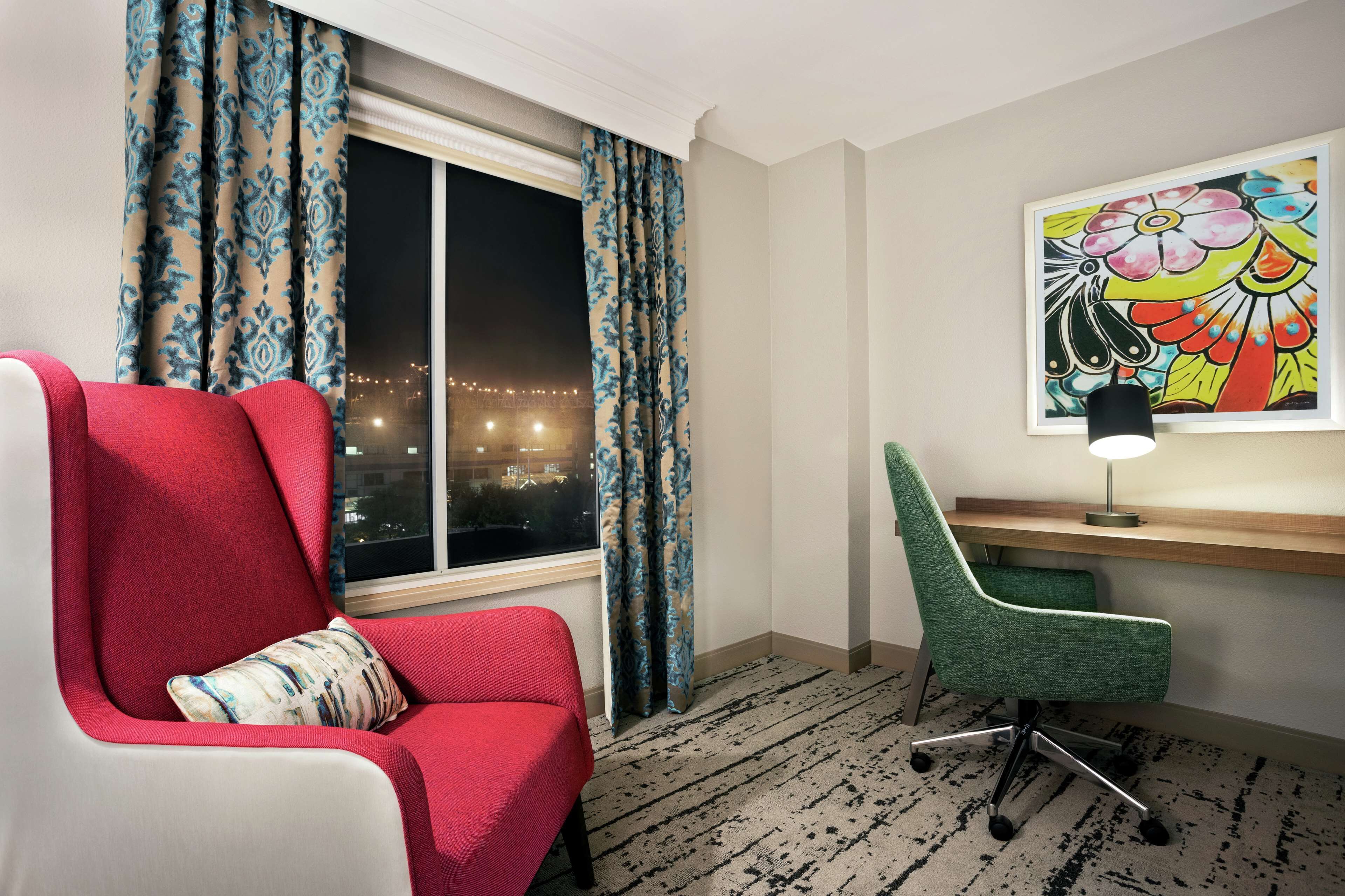Una habitación de hotel con un sillón rojo, una silla de escritorio verde, un escritorio con lámpara, una ventana con cortinas y un cuadro de colores en la pared.