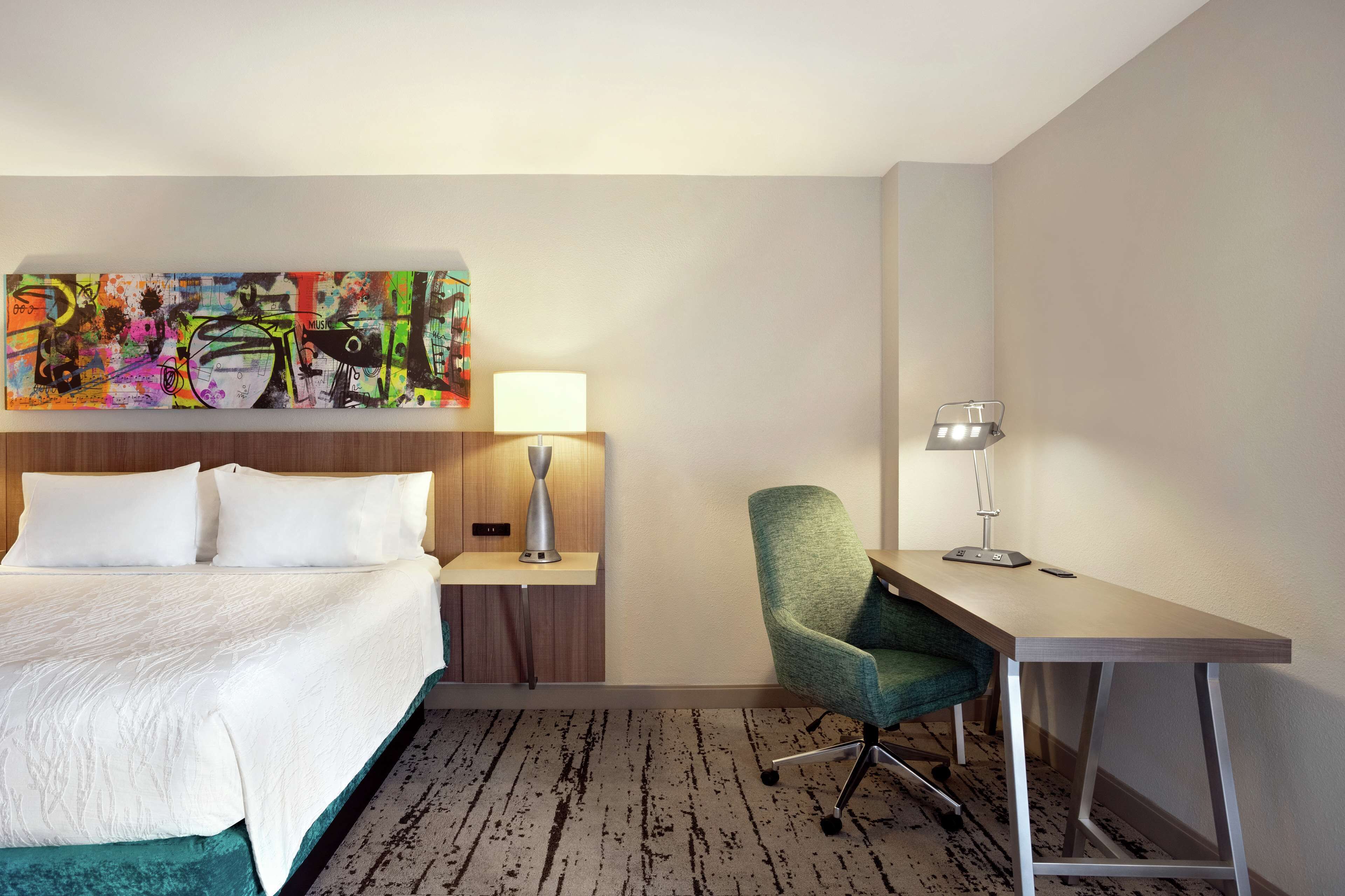 A hotel room with a bed, a desk, a chair, a lamp, and a colorful abstract painting on the wall.