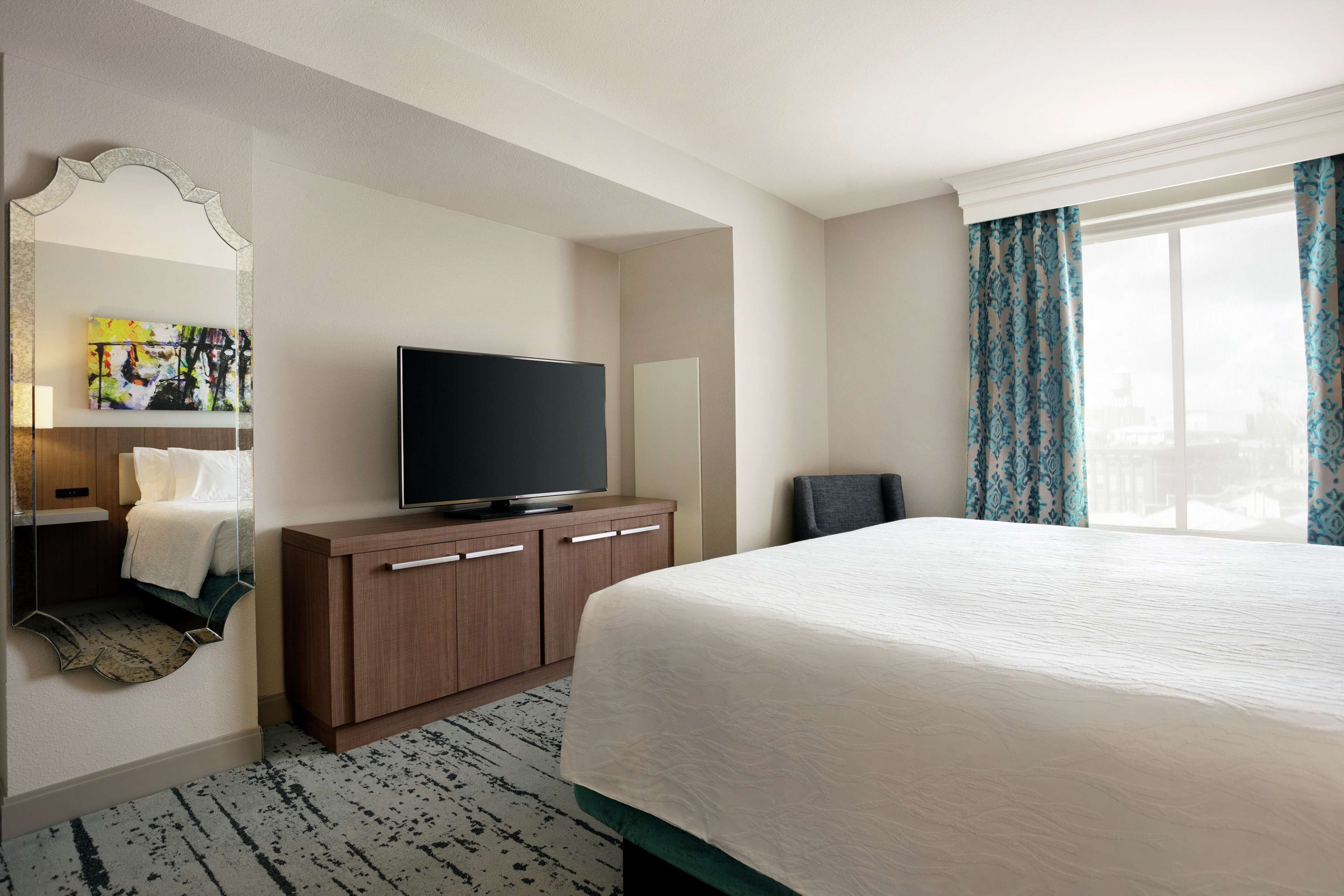 A hotel room with a large bed, a flat-screen TV, a wooden dresser, a mirror, and a window with curtains.