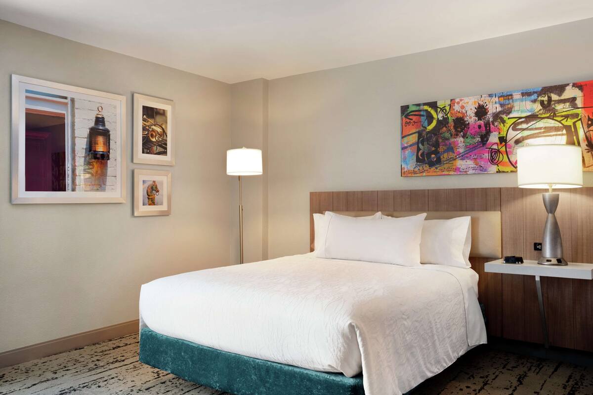 A hotel room with a large bed, a wooden headboard, a nightstand with a lamp, and three framed pictures on the wall.