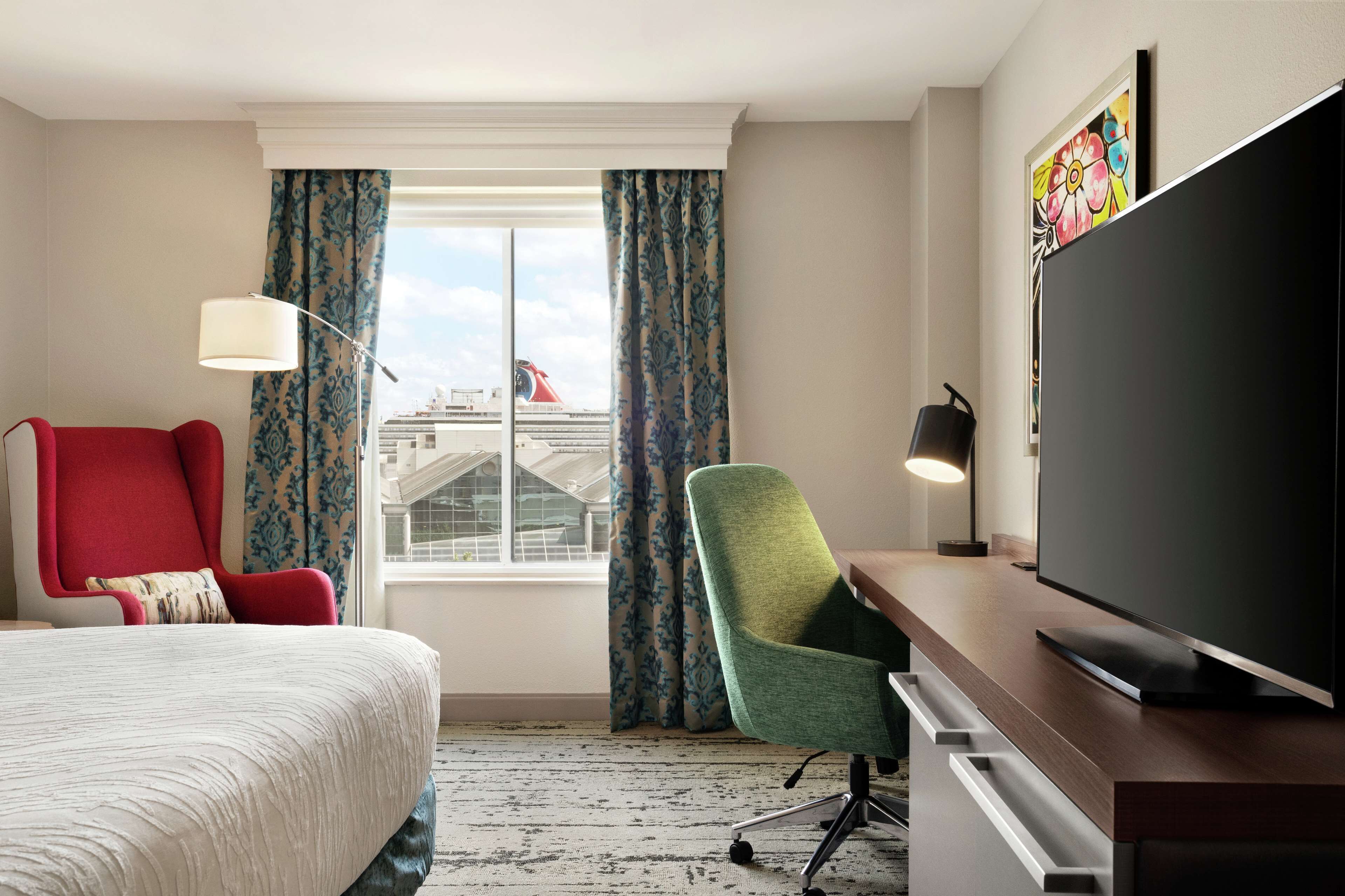 Habitación de hotel con cama, escritorio con televisor de pantalla plana, silla de oficina verde, sillón rojo y ventana con cortinas.