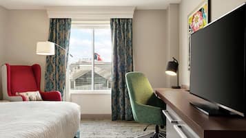 Habitación de hotel con cama, escritorio con televisor de pantalla plana, silla de oficina verde, sillón rojo y ventana con cortinas.