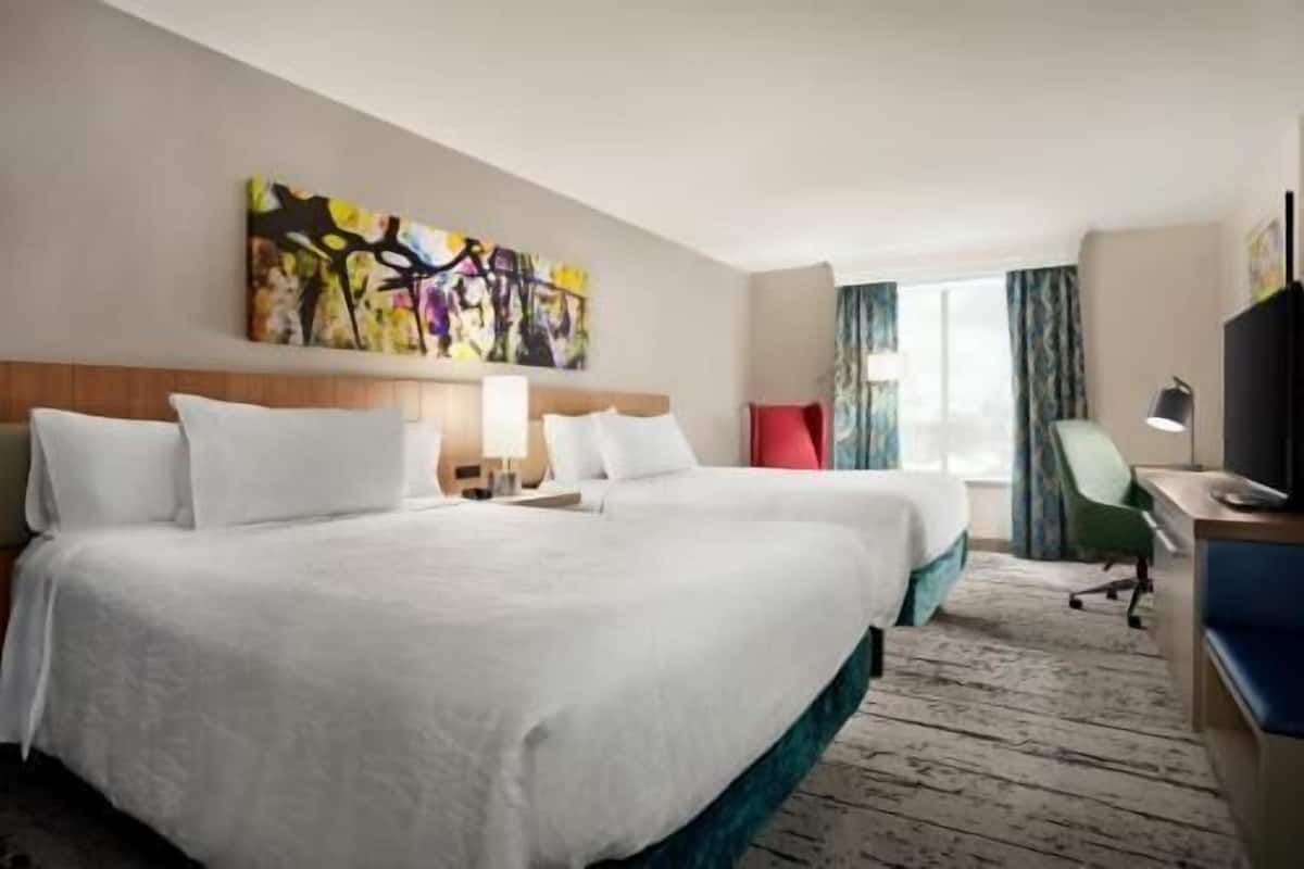 A hotel room with two beds, a desk, a chair, a TV, and a painting on the wall.