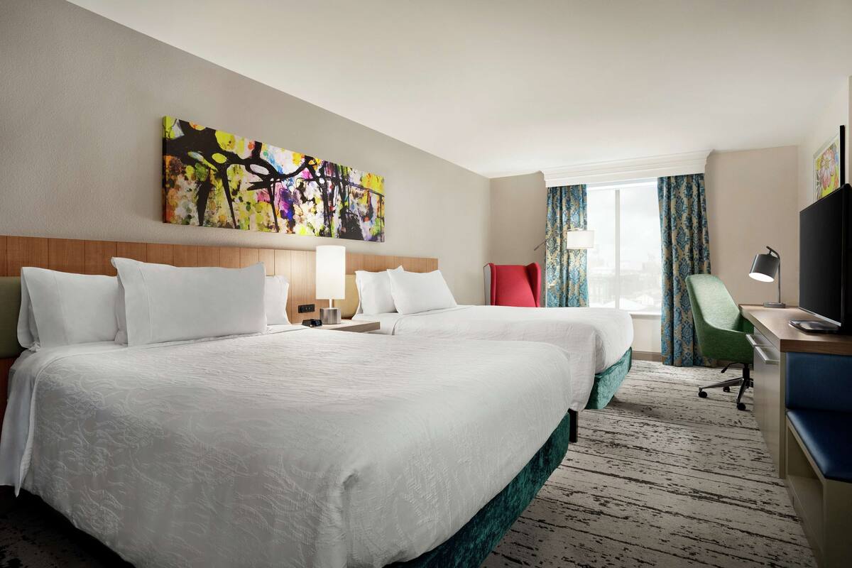 A hotel room with two beds, a desk, a chair, a TV, and a window with curtains.