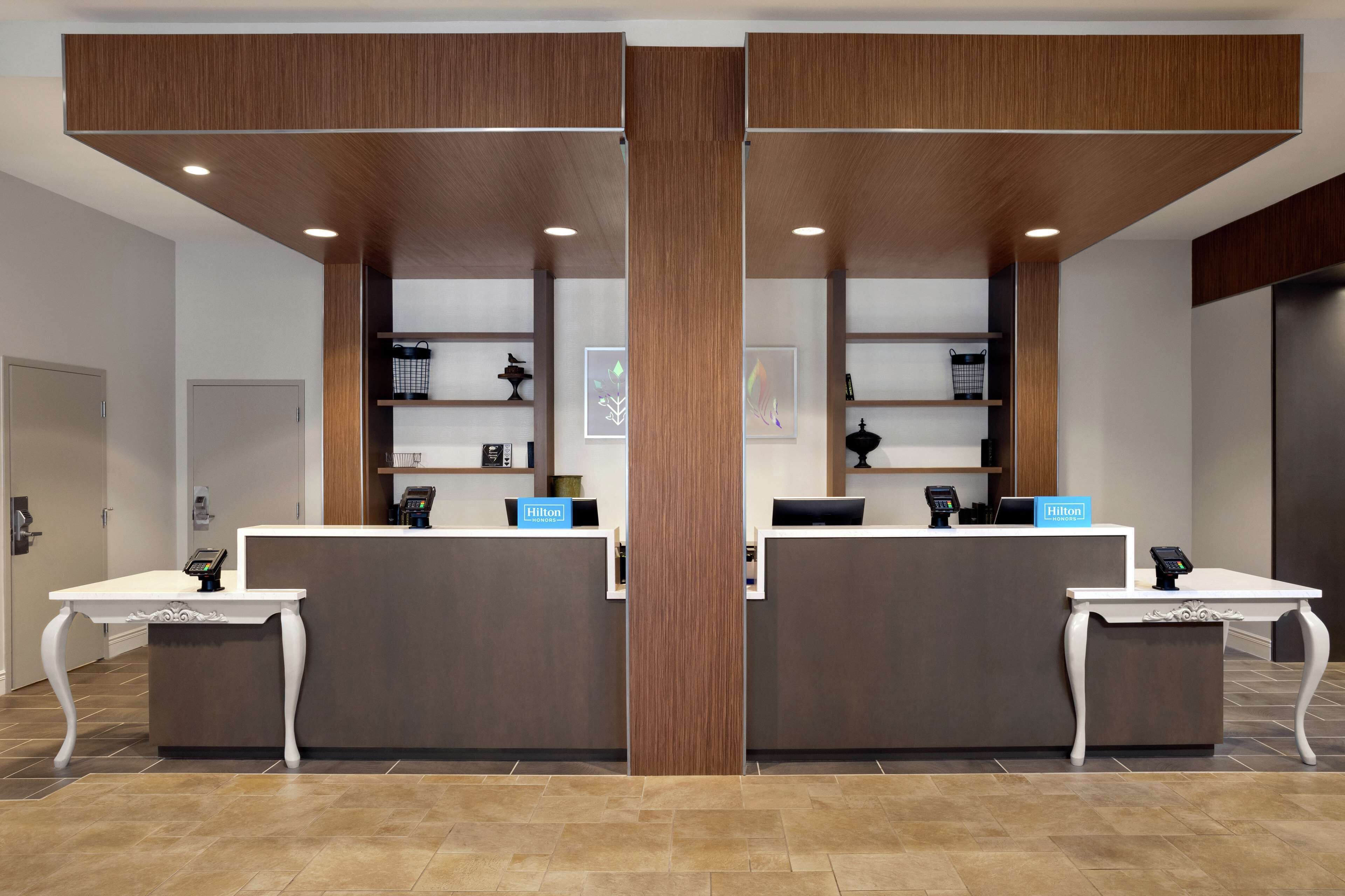 A hotel reception area with two counters, each with a digital kiosk and a telephone. There are shelves with various items and a large wooden panel above the counters.