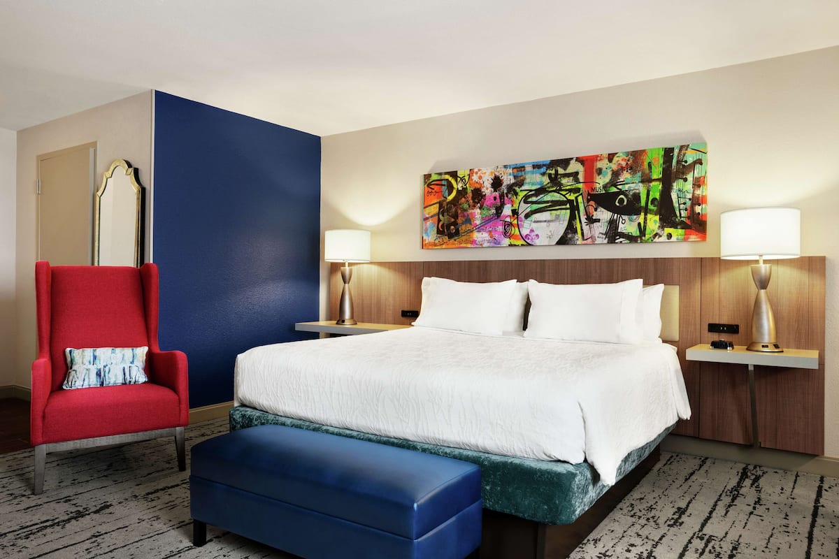 A hotel room with a large bed, a red armchair, a blue bench, two bedside tables with lamps, and a colorful abstract painting on the wall.