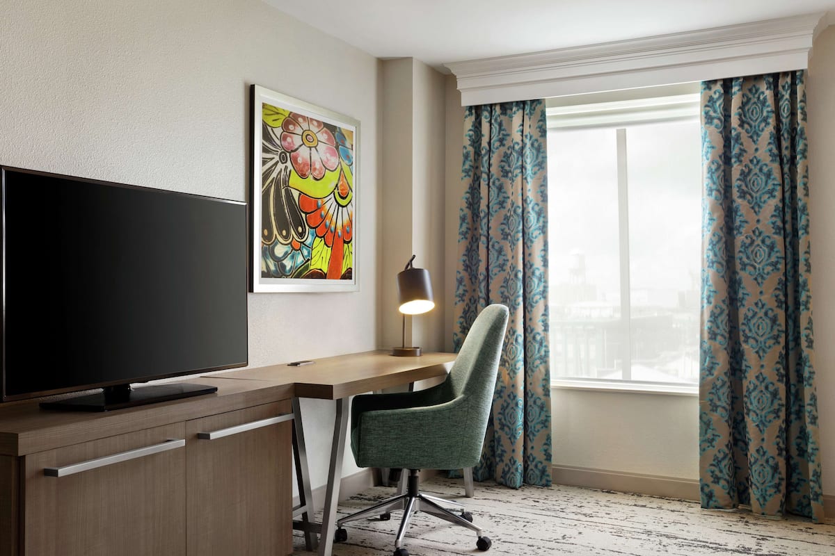 A modern hotel room with a flat-screen TV, a desk, a chair, and a window with patterned curtains.