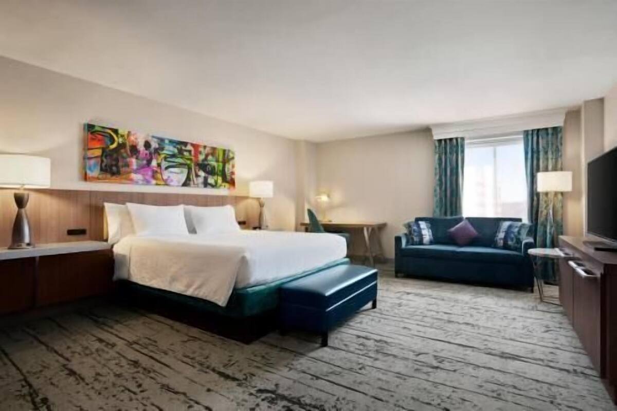 A hotel room with a bed, a sofa, a desk, a TV, and a painting on the wall.