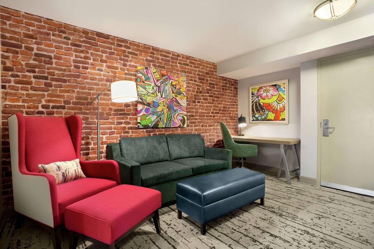 A room with a brick wall, a green sofa, a red armchair, a blue ottoman, and a desk with a lamp.