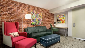 A room with a brick wall, a green sofa, a red armchair, a blue ottoman, and a desk with a lamp.