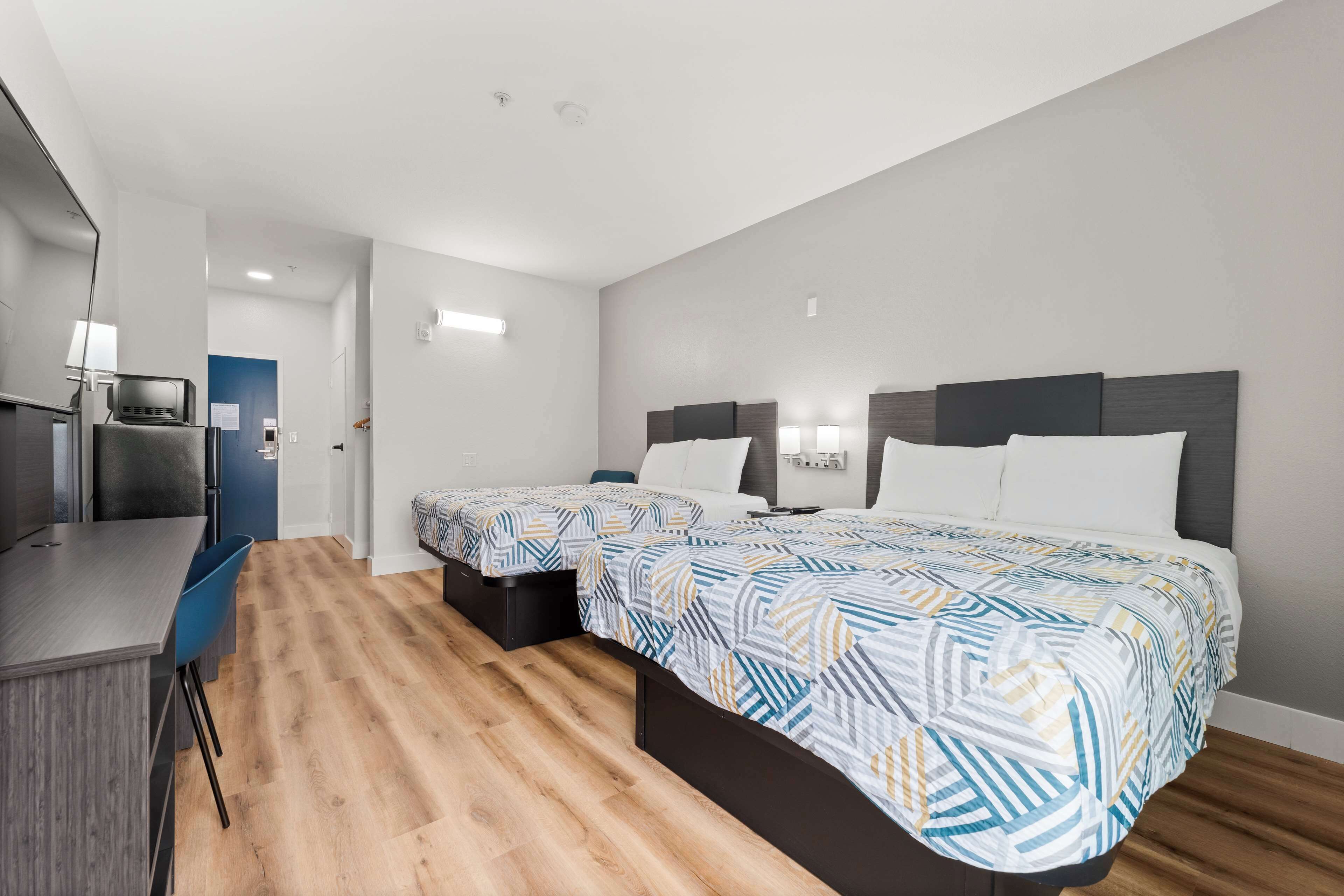 A modern hotel room with two single beds, a small desk, and a kitchenette.