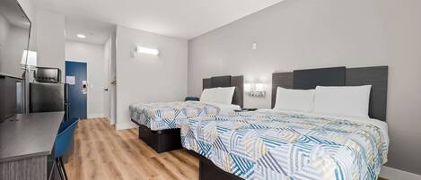 A modern hotel room with two single beds, a small desk, and a kitchenette.