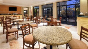Un amplio comedor interior con varias mesas y sillas redondas, una zona de cocina al fondo y grandes ventanales que dejan entrar luz natural.