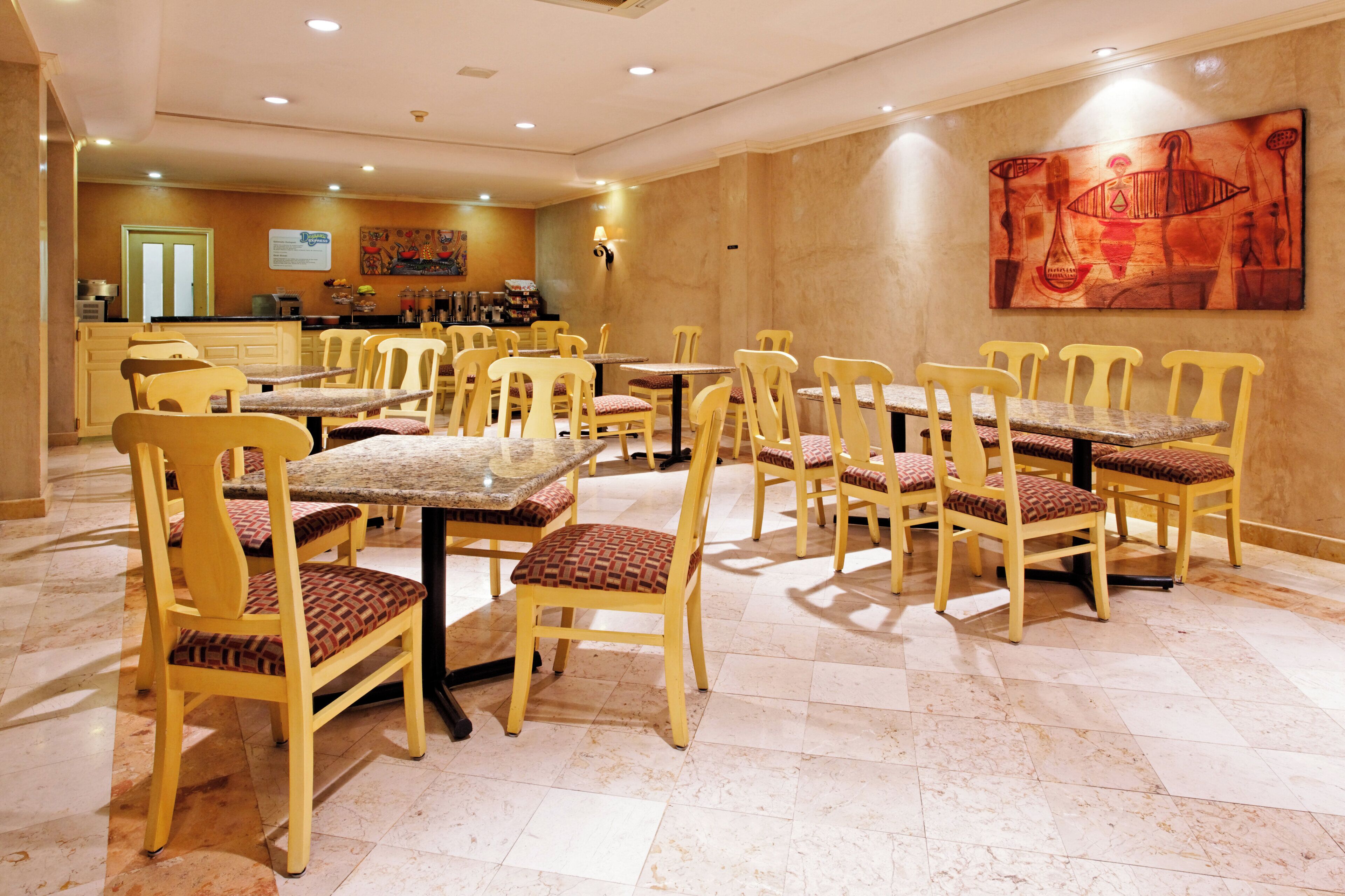 A hotel dining area with multiple tables and chairs, a kitchen in the background, and a large painting on the wall.