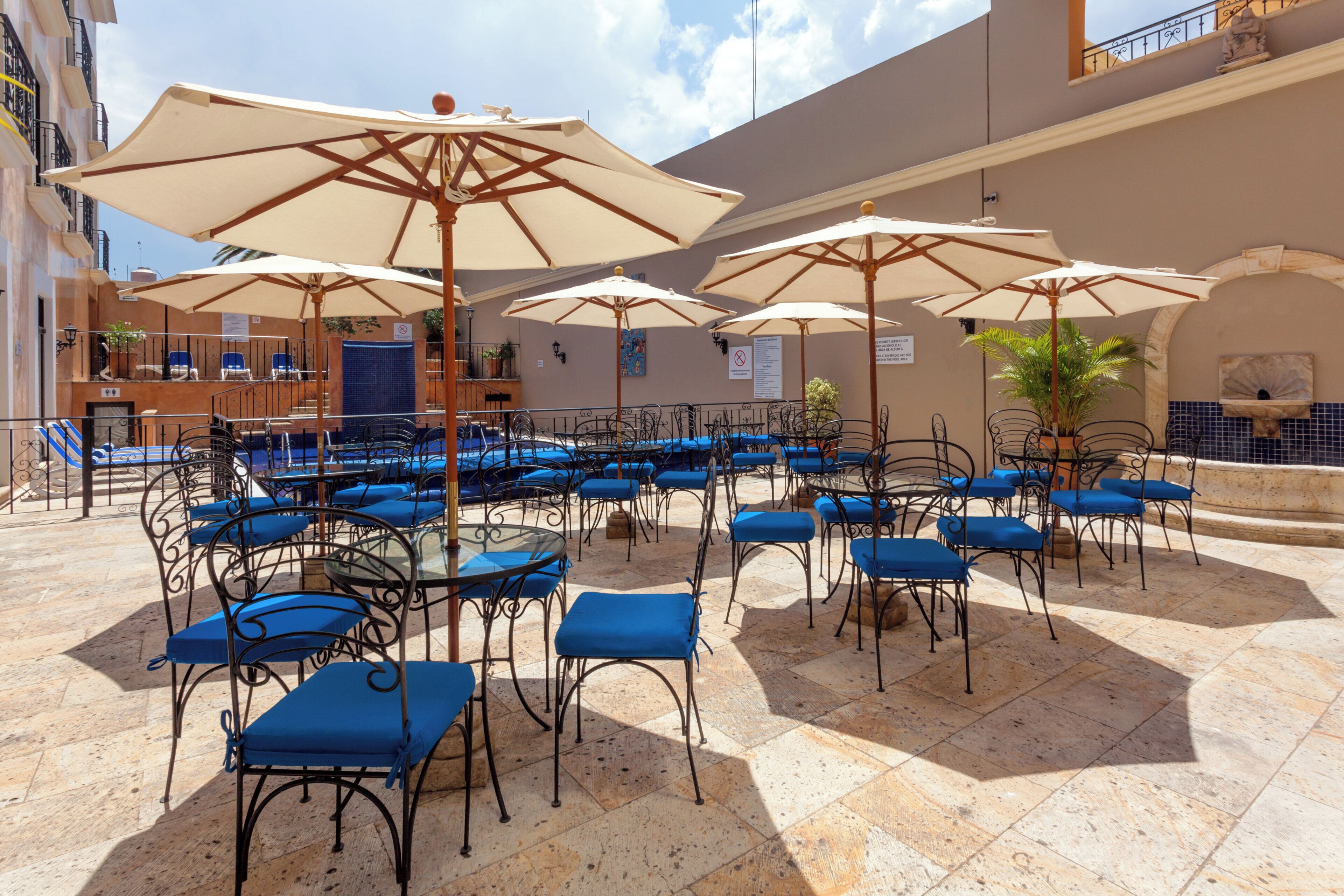 An outdoor seating area with multiple tables and chairs, umbrellas, and a pool in the background.