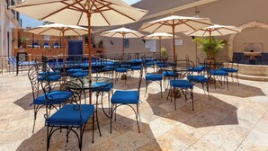 Un espacio al aire libre con varias mesas y sillas, sombrillas y una piscina al fondo.