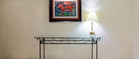 A framed artwork above a glass-top console table with a lamp beside it.