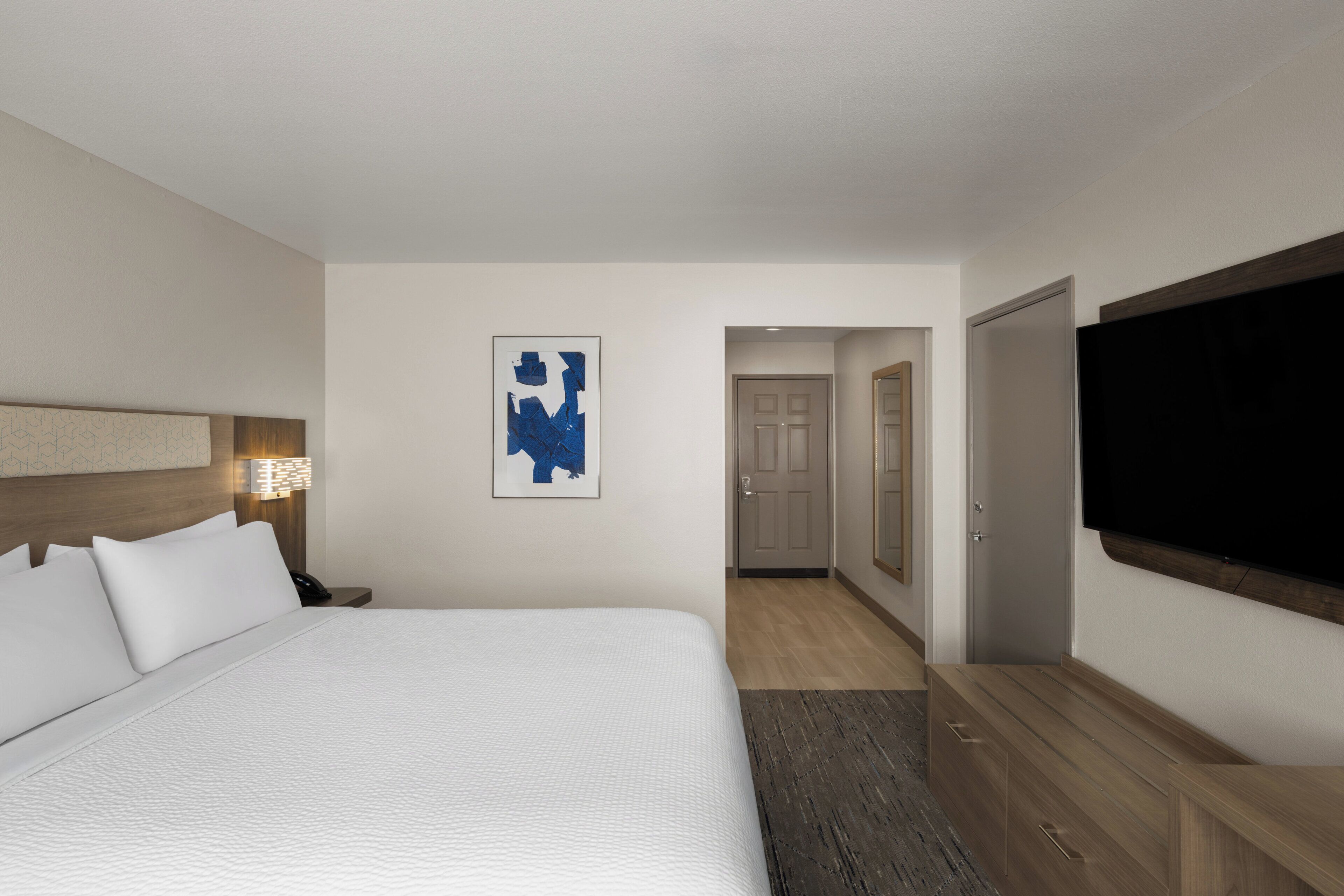 A hotel room with a large bed, a flat-screen TV, a framed abstract painting, and a wooden headboard.