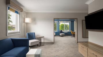A hotel room with a blue sofa, a small round table, a TV mounted on the wall, and a view through an open door to a balcony area.
