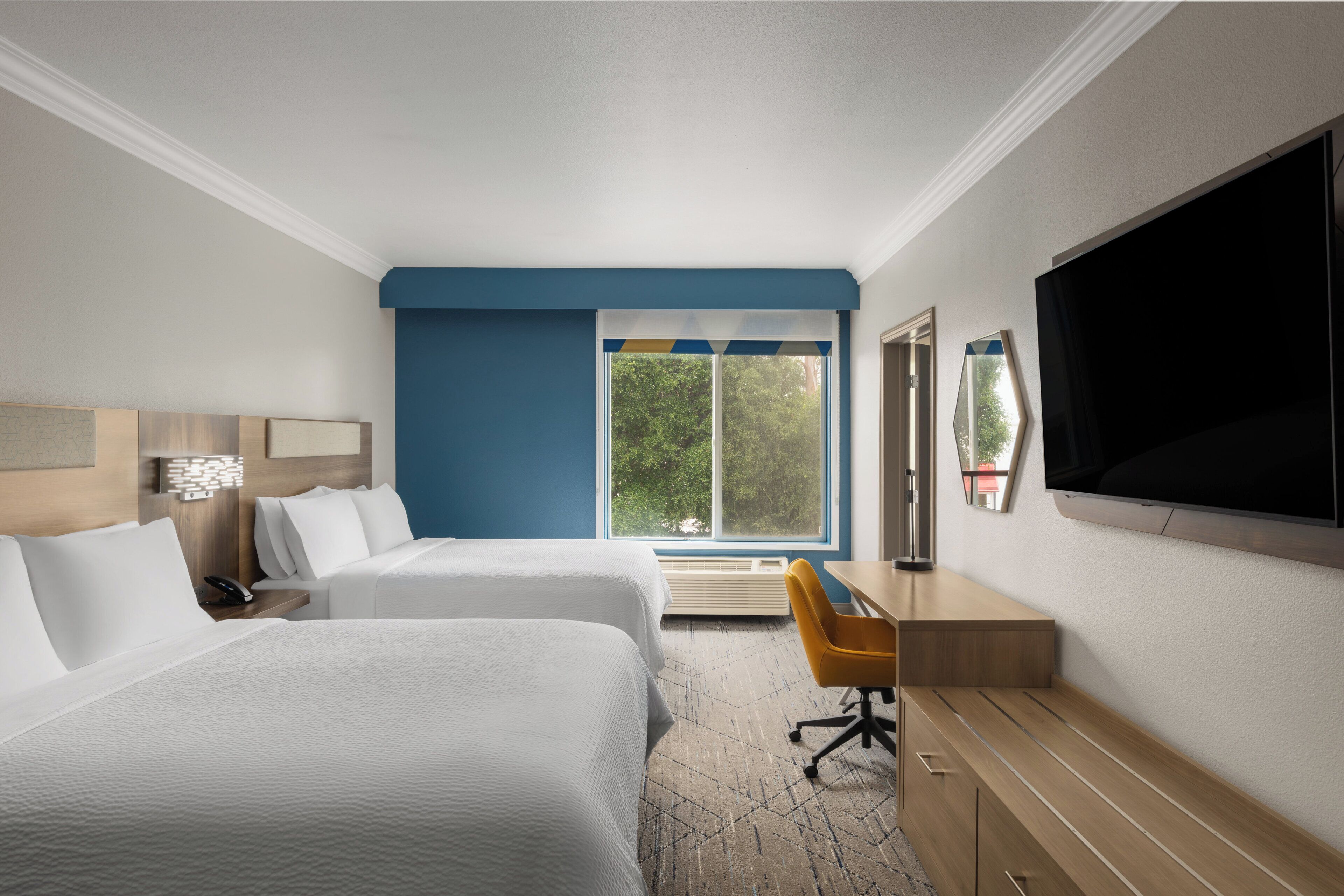 Habitación de hotel con dos camas, un escritorio con silla, un televisor de pantalla plana y un ventanal con vistas a árboles.